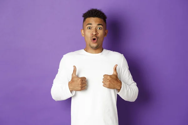 Impressed handsome african-american guy, showing thumbs-up in approval, praise great promo, like and recommend, looking amazed at camera, standing over purple background — Stock Photo, Image