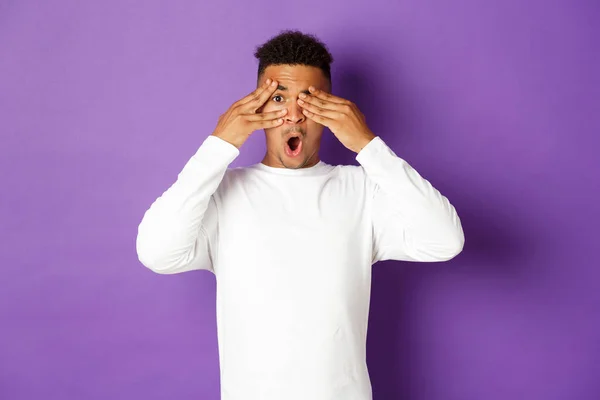 Retrato de curioso homem afro-americano, à espera de surpresa, olhos fechados com as mãos, mas espreitando através dos dedos em algo interessante, de pé sobre fundo roxo — Fotografia de Stock