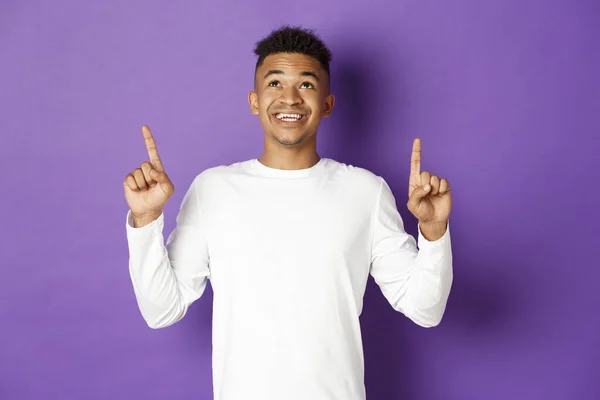 Cheerful african-american man in white sweatshirt, showing logo, pointing and looking up with happy smile, standing over purple background — Stock Photo, Image