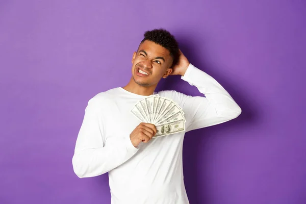 Imagen de hombre afroamericano confuso, sosteniendo efectivo y mirando la esquina superior izquierda desconcertado, pensando qué comprar, de pie sobre fondo púrpura — Foto de Stock
