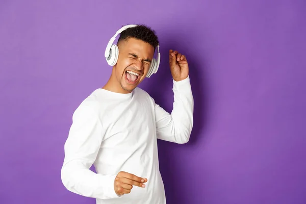 Imagen de un chico afroamericano despreocupado bailando, escuchando música en auriculares inalámbricos y cantando, de pie sobre fondo púrpura —  Fotos de Stock