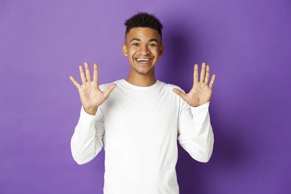 Image of cheerful african-american guy in white sweatshirt, showing number ten and smiling, standing over purple background — Stock Photo, Image