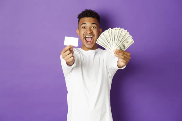 Imagen de chico afroamericano guapo, mirando complacido y sonriente, mostrando tarjeta de crédito y dinero, de pie sobre fondo púrpura — Foto de Stock