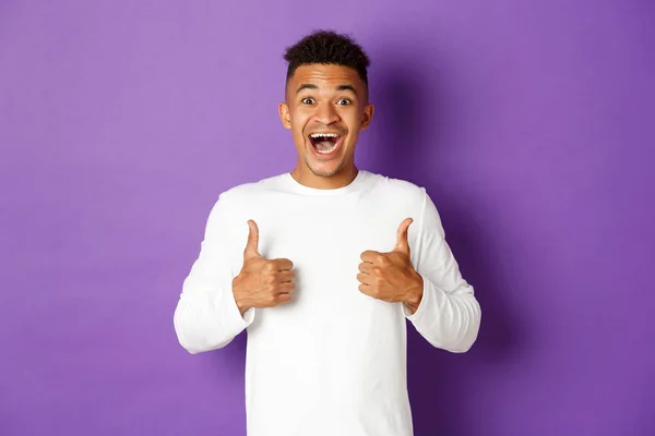 Excited african-american man showing thumbs-up, smiling amazed, like and praise great promo, standing over purple background — Stock Photo, Image