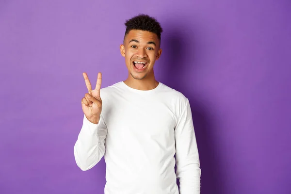 Image d'un homme afro-américain joyeux en sweat-shirt blanc, montrant deux doigts et souriant, debout sur un fond violet — Photo