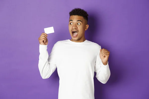 紫の背景の上に立って、驚きとクレジットカードを見て、印象的なアフリカ系アメリカ人の男 — ストック写真