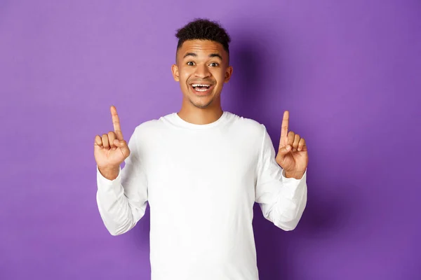 Alegre cara afro-americano em camisola branca, apontando os dedos para cima e sorrindo, fazendo um anúncio, mostrando seu logotipo no espaço de cópia, de pé sobre fundo roxo — Fotografia de Stock