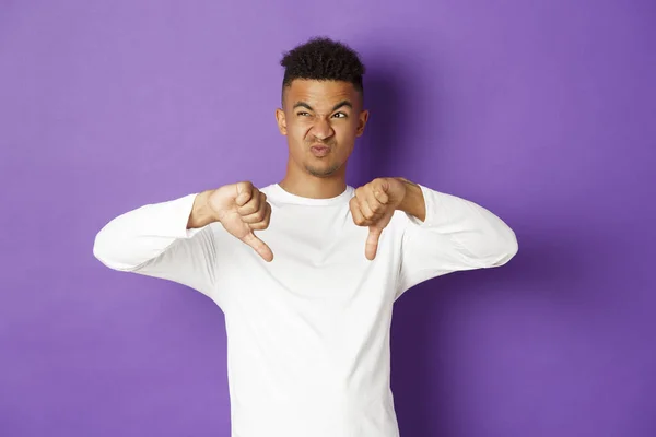 Image of skeptical and disappointed african-american young man, looking at something disgusting at upper left corner, showing thumbs-down and grimacing from dislike, standing over purple background — Stock Photo, Image