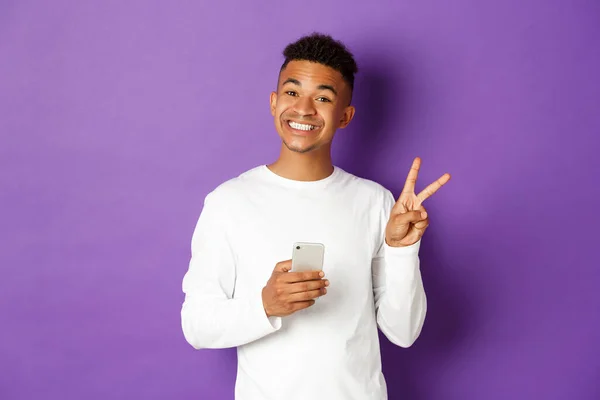 Bild eines gut aussehenden afrikanisch-amerikanischen Mannes, der lächelt und ein Friedenszeichen zeigt, während er sein Mobiltelefon benutzt, vor lila Hintergrund — Stockfoto