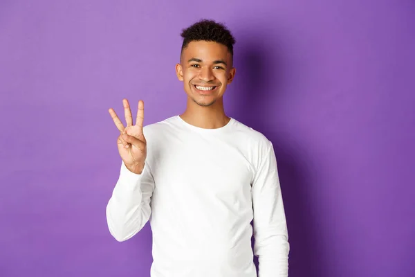 Bild von fröhlichen afrikanisch-amerikanischen Kerl in weißem Sweatshirt, zeigt drei Finger und lächelt, steht über lila Hintergrund — Stockfoto