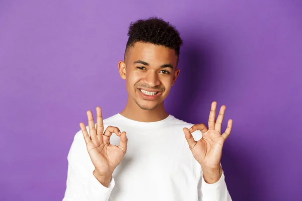 Крупный план молодого афро-американского парня, выглядящего довольным, улыбающимся и показывающим нормальные признаки, одобряющего и любящего что-то хорошее, стоящего на фиолетовом фоне — стоковое фото