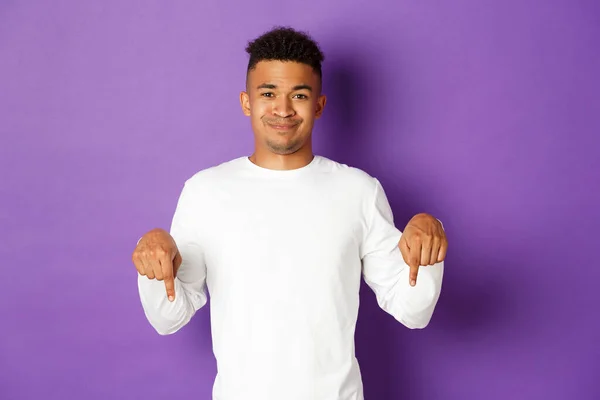 Image d'un homme afro-américain sceptique en sweat-shirt blanc, déçu, souriant et pointant du doigt quelque chose de désagréable, debout sur un fond violet — Photo