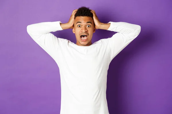 Image d'un bel homme afro-américain paniqué, tenant la tête et regardant la caméra nerveuse, alarmé et effrayé sur fond violet — Photo