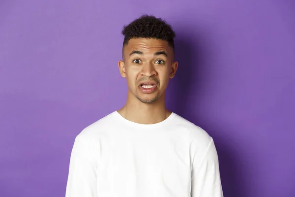 Close-up of startled african-american man in white sweatshirt, gasping and open mouth in awe, standing over purple background — Stock Photo, Image