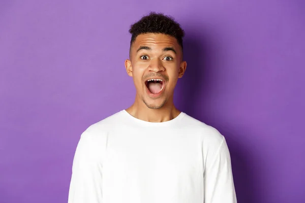 Close-up of amazed african-american young man, open mouth and saying wow, looking fascinated at camera, standing over purple background with copy space — Stock Photo, Image