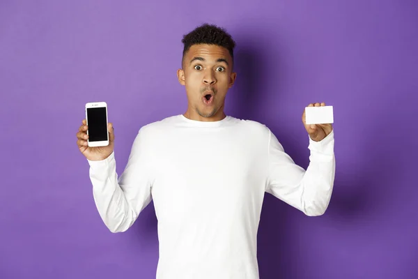 Портрет красивого африканско-американского мужчины, выглядящего изумленным, показывающего экран смартфона и кредитную карту, стоящего на фиолетовом фоне — стоковое фото
