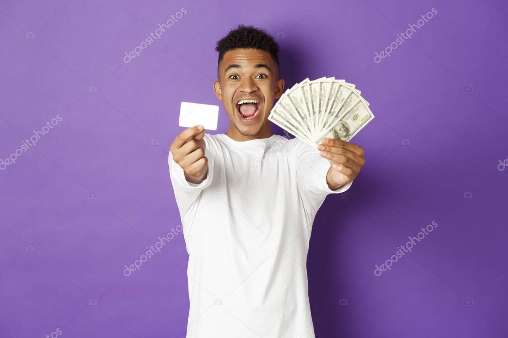 Image of handsome african american guy, looking pleased and smiling, showing credit card and money, standing over purple background