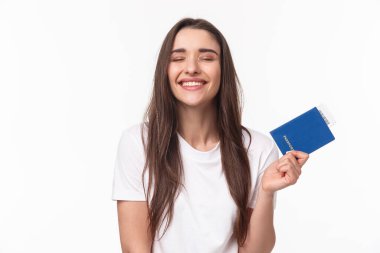 Seyahat, tatil, yaz konsepti. Beyaz tişörtlü mutlu genç kız, gözleri kapalı gülümseyen, tatilde kumsalda güneşlendiğini hayal eden, pasaportunu biletle tutan.