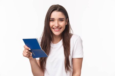 Seyahat, tatil, yaz konsepti. Güzel, mutlu, tişörtlü, elinde açık pasaport ve gülümsemeyle havaalanı terminalinde duran seyahate çıkan genç bir kadının yakın plan portresi.