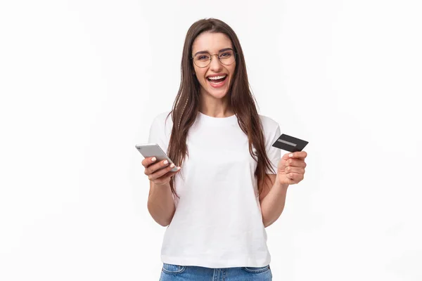 Retrato de mujer bonita sonriente feliz en gafas con tarjeta de crédito, teléfono móvil, riéndose de la cámara pagando fácilmente por su tienda en línea de compra, comprar cosas con aplicación, fondo blanco — Foto de Stock