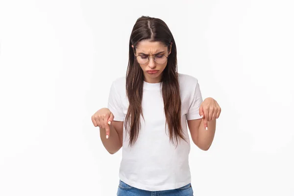 Waist-up portrait of unsure, skeptical young brunette female in glasses and t-shirt, look perplexed, cant figure out what is this lying down, pointing and looking bottom advertisement puzzled — Stock Photo, Image