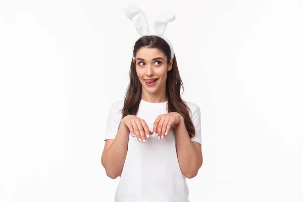 Rolig och dum, lekfull flicka i kaninöron, t-shirt, tittar bort och slickar läppar, dagdrömmer om något läckert på påskdagen, imitera kanin med händerna dras nära bröstet som tassar — Stockfoto