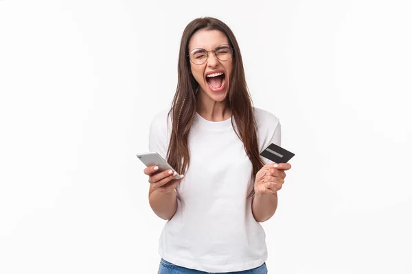 Portrait of uneasy annoyed and angry young brunette female in t-shirt yelling pissed-off as checking bank account, have no money on credit card, or it expired, hold mobile phone, white background — Foto Stock