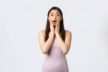 Kutlama, güzellik ve cazibe konsepti. Şok olmuş ve ürkmüş genç Asyalı kadın lüks bir gece elbisesi içinde, çenesini indir ve şaşırmış kameraya bak, beyaz arka plan