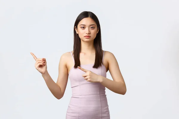 Concepto de celebración, belleza y glamour. Mujer asiática de aspecto serio en elegante vestido de noche, mostrando dónde fiesta, camino al evento de vacaciones, señalando los dedos a la izquierda, fondo blanco —  Fotos de Stock