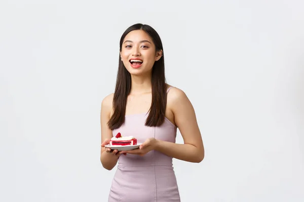 Lifestyle, feestdagen, feesten en eten concept. Enthousiast mooi aziatisch meisje eten op feest, het dragen van jurk, holding plaat met stuk taart, lachen en glimlachen, witte achtergrond — Stockfoto