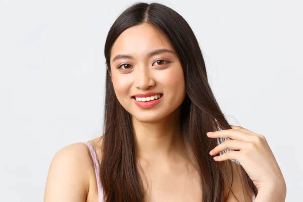 Schoonheid, mode en mensen emotie concept. Close-up portret van mooie kokette aziatische vrouw met vrolijke glimlach, aanraken van haar en staren camera romantisch, witte achtergrond — Stockfoto