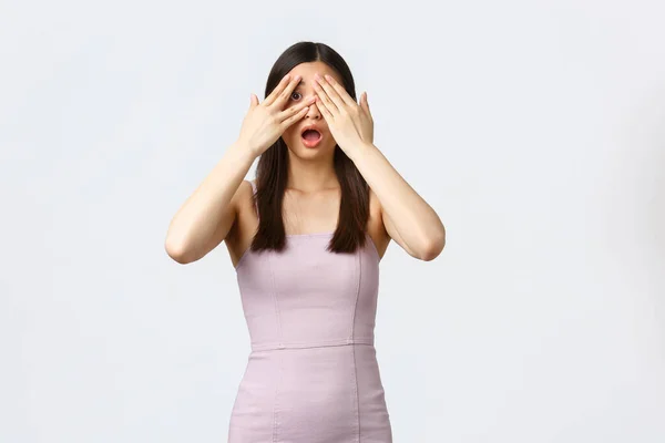 Mujeres de lujo, fiesta y concepto de vacaciones. Emocionado chica de cumpleaños asiática atractiva en vestido elegante, ojos cerrados con palmas y mirar a través de los dedos, a la espera de regalo sorpresa, fondo blanco — Foto de Stock