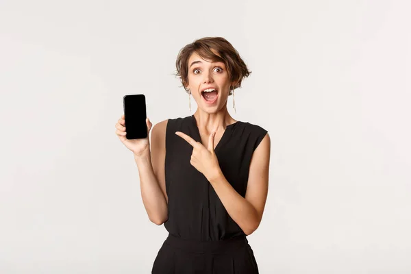 Úžasné atraktivní dívka vypadá vzrušeně, ukazuje prstem na displej smartphone, stojící bílé pozadí — Stock fotografie