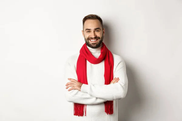 Vacaciones de Navidad y concepto de celebración. Hombre barbudo guapo en suéter celebrando año nuevo, vistiendo bufanda roja y sonriendo, de pie sobre fondo blanco — Foto de Stock