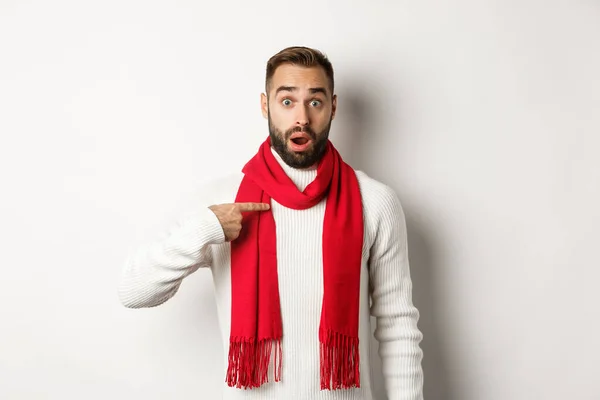 Zimní dovolená a nákupní koncept. Překvapený a zmatený chlap ukazuje na sebe, je vybrán, stojí v červené šále a svetru na bílém pozadí — Stock fotografie