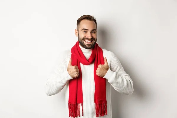 Winterurlaub und Einkaufskonzept. Fröhlicher bärtiger Mann in rotem Weihnachtsschal zeigt Daumen hoch, genehmigt und empfiehlt Laden, steht über weißem Hintergrund — Stockfoto