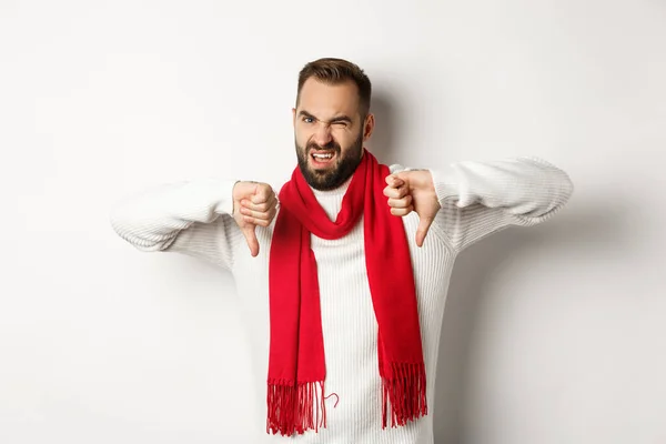 Skeptischer und unzufriedener Mann zeigt Daumen nach unten, trägt Winterpullover mit Schal, widerspricht, steht über weißem Hintergrund — Stockfoto