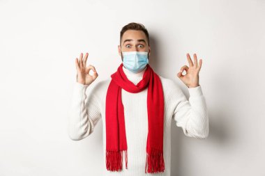 Covid-19, tecrit ve karantina konsepti. Yüz maskeli etkileyici bir adam, tamam işaretleri gösteriyor ve katılıyor, harika bir ürün öneriyor, beyaz arka plan.