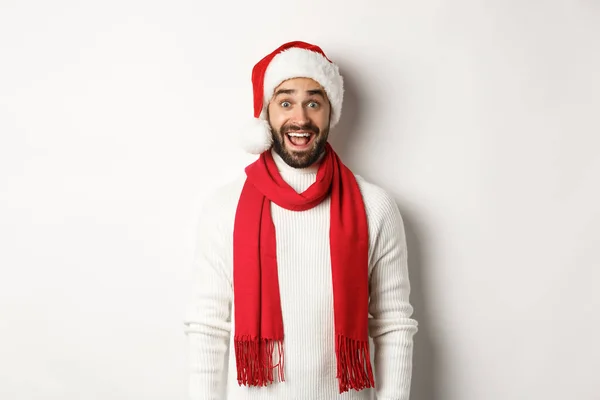 Férias de Natal. Homem barbudo olhando surpreso para a câmera, vestindo chapéu de festa de Papai Noel e cachecol vermelho, de pé contra fundo branco — Fotografia de Stock