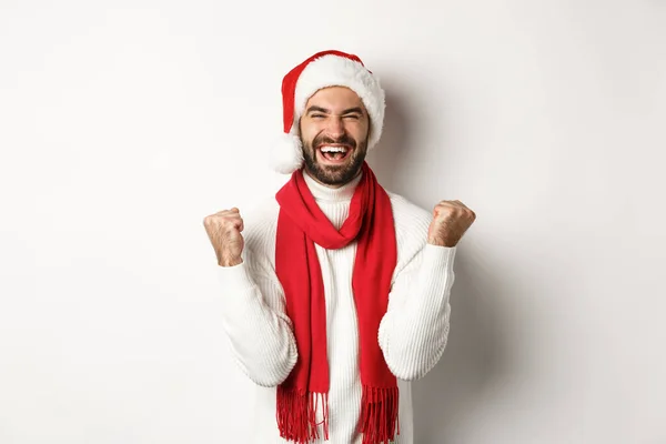 Christmas holidays. Man winner celebrating and triumphing, raising hands up in rejoice, achieve goal, wearing Santa hat and red scarf, white background — Stock Photo, Image