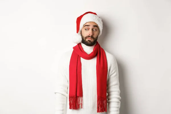 Férias de inverno e conceito de festa de Ano Novo. Cara curiosa espreitando no banner do logotipo esquerdo, de pé no chapéu de Papai Noel, celebrando o Natal, de pé sobre fundo branco — Fotografia de Stock