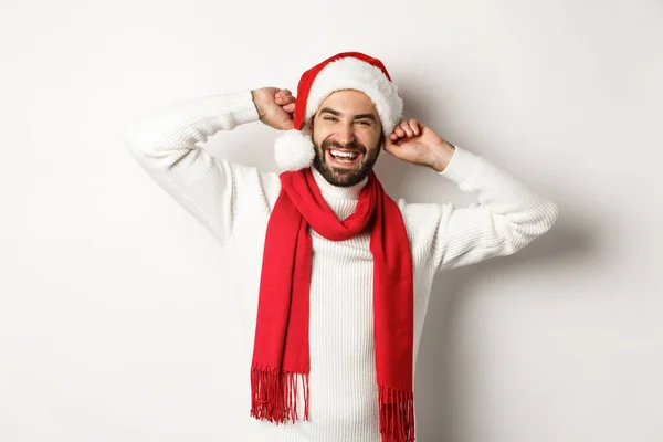 Winterurlaub und Neujahrsparty. Glücklicher Mann feiert Weihnachten, trägt Weihnachtsmütze und roten Schal, lacht laut, weißer Hintergrund — Stockfoto