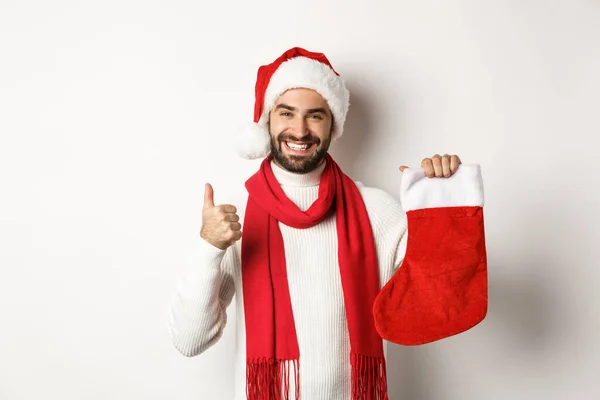 Férias de inverno e conceito de celebração. Homem feliz mostrando meia de Natal para presentes e polegar para cima, sorrindo satisfeito, de pé sobre fundo branco — Fotografia de Stock