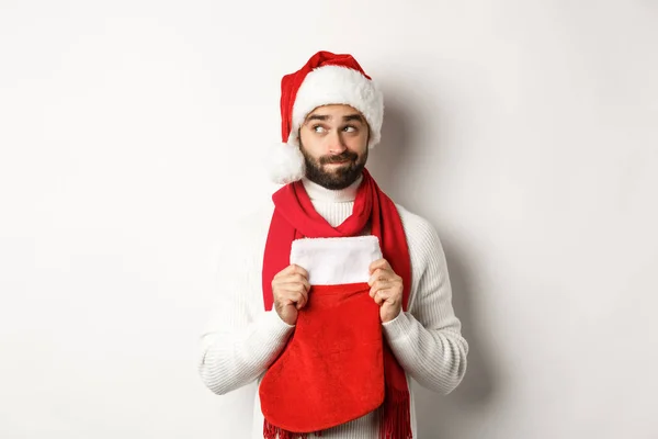 Zimní dovolená a nákupní koncept. Hezký vousatý chlap v Santa klobouk přemýšlet o dárky, držení vánoční ponožky, stojící nad bílým pozadím — Stock fotografie