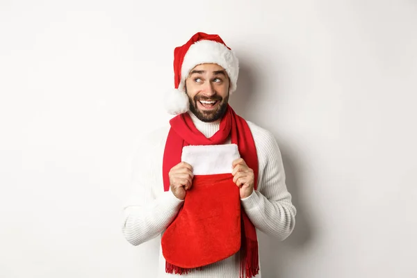 Zimowe wakacje i koncepcja zakupów. Podekscytowany przystojny mężczyzna planuje nowy rok, trzymając skarpetki świąteczne na prezenty i myślenia, stojąc na białym tle — Zdjęcie stockowe