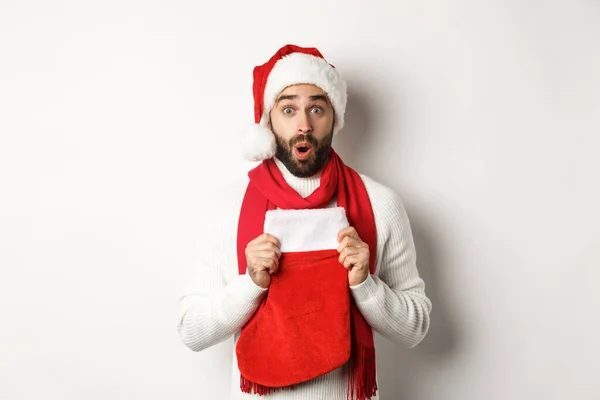 Winterurlaub und Einkaufskonzept. Überraschter Mann mit Weihnachtsmütze erhält Geschenk in Weihnachtssocke, schaut erstaunt, steht vor weißem Hintergrund — Stockfoto