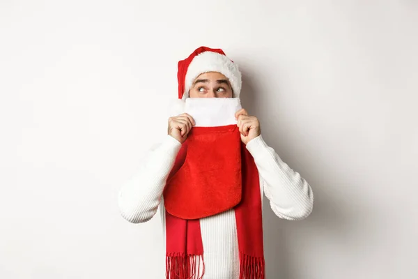 Vintersemester och shopping koncept. Glada kaukasiska killen i Santa hatt gömmer ansiktet bakom julstrumpa med gåvor, stående över vit bakgrund — Stockfoto