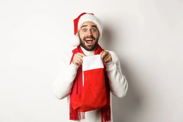 Зимние праздники и праздничная концепция. Удивленный бородатый мужчина смотрит в рождественский носок, получает новогодний подарок, стоит на белом фоне — стоковое фото