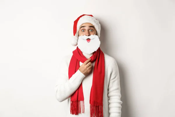 Julfest och firande koncept. Rolig ung man i Santa hatt håller vitt skägg mask och göra ansikten, njuta av nyår, vit bakgrund — Stockfoto