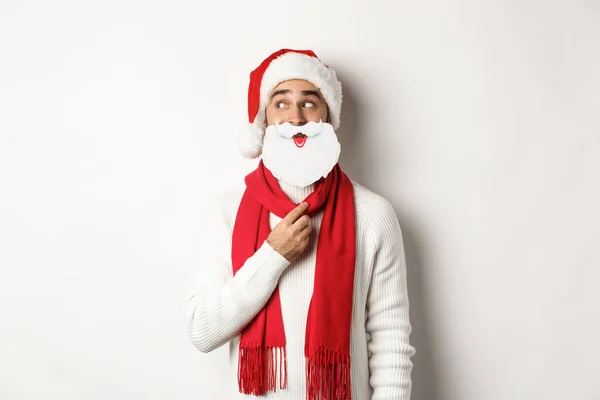 Festa de Natal e conceito de celebração. Homem engraçado desfrutando de Ano Novo, segurando máscara de barba branca de Santa e vestindo chapéu vermelho, fundo de estúdio — Fotografia de Stock